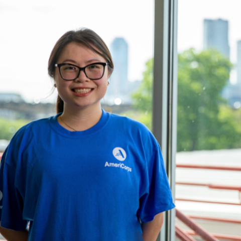 Woman wearing an AmeriCorps T-shirt and standing in front of a large glass window with bright greenery outside